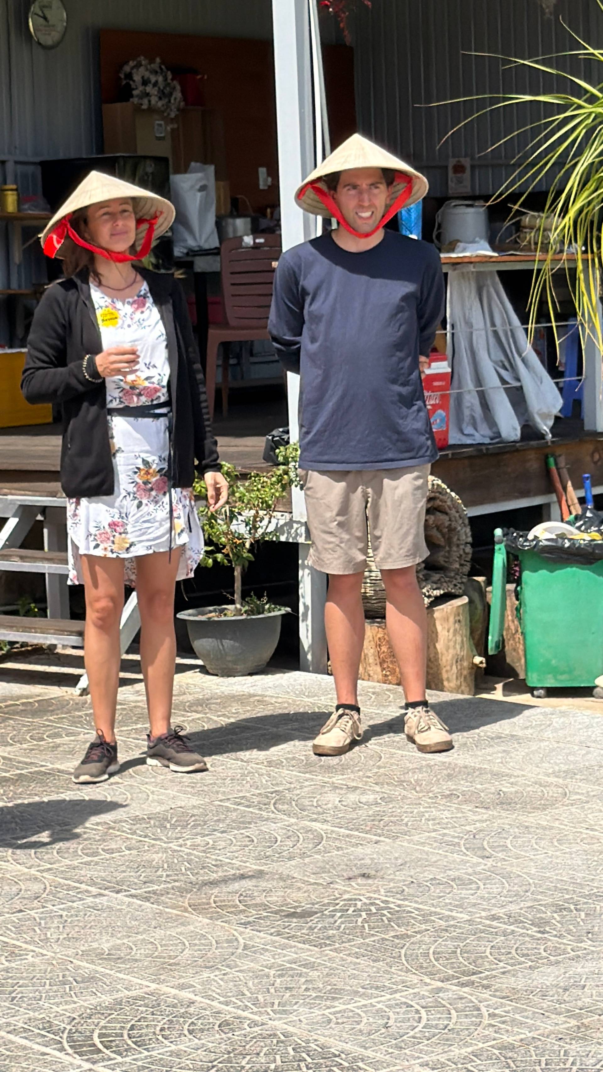 Two visitors wearing traditional Vietnamese conical hats at an outdoor community event venue in Đà Lạt, set against a rustic workshop backdrop with tropical plants and decorative elements.