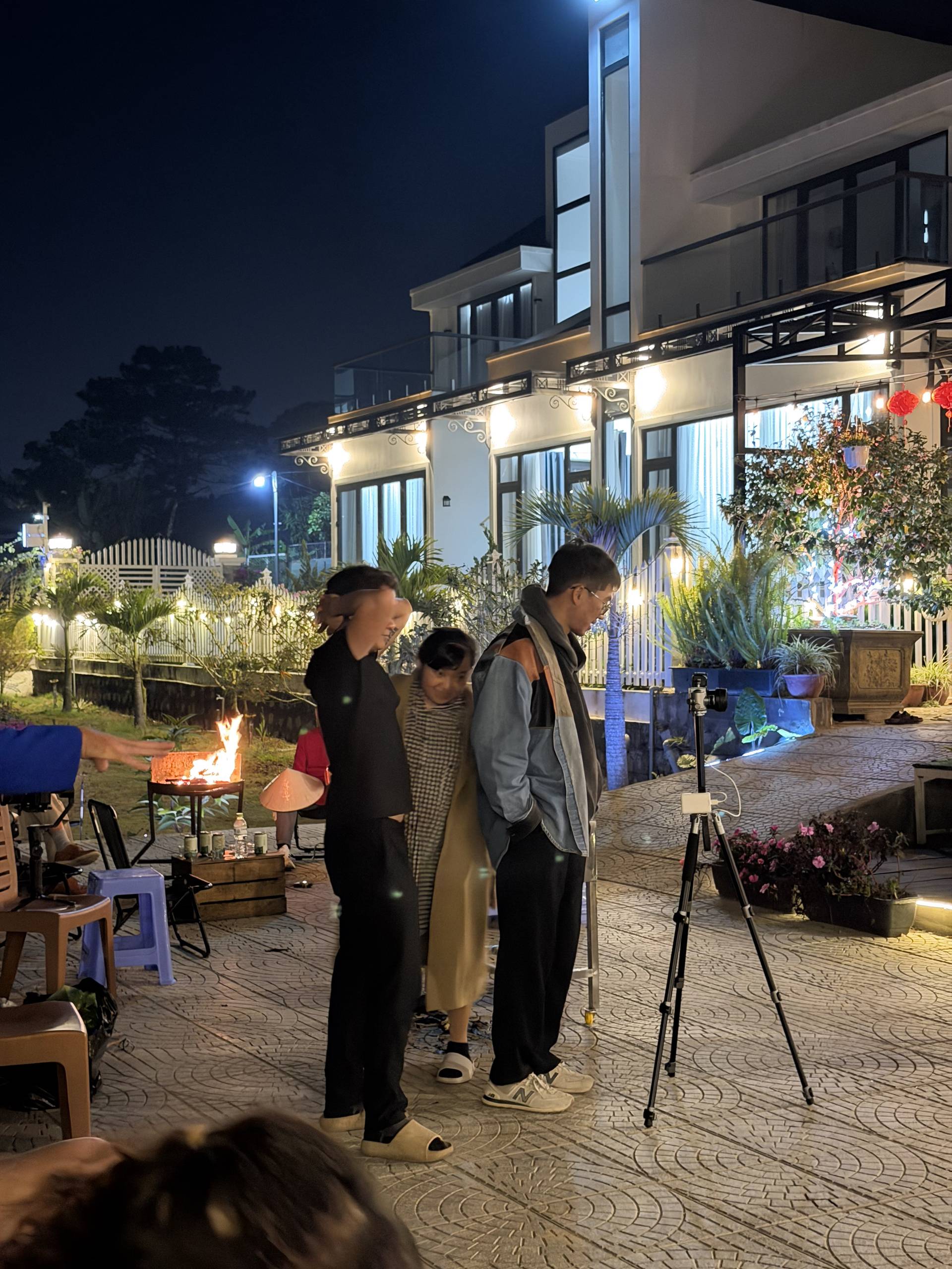 Evening social gathering at an elegant modern villa in Đà Lạt with warm ambient lighting and outdoor fire pit. Guests mingle on a decorated paved courtyard surrounded by tropical plants and contemporary architecture.