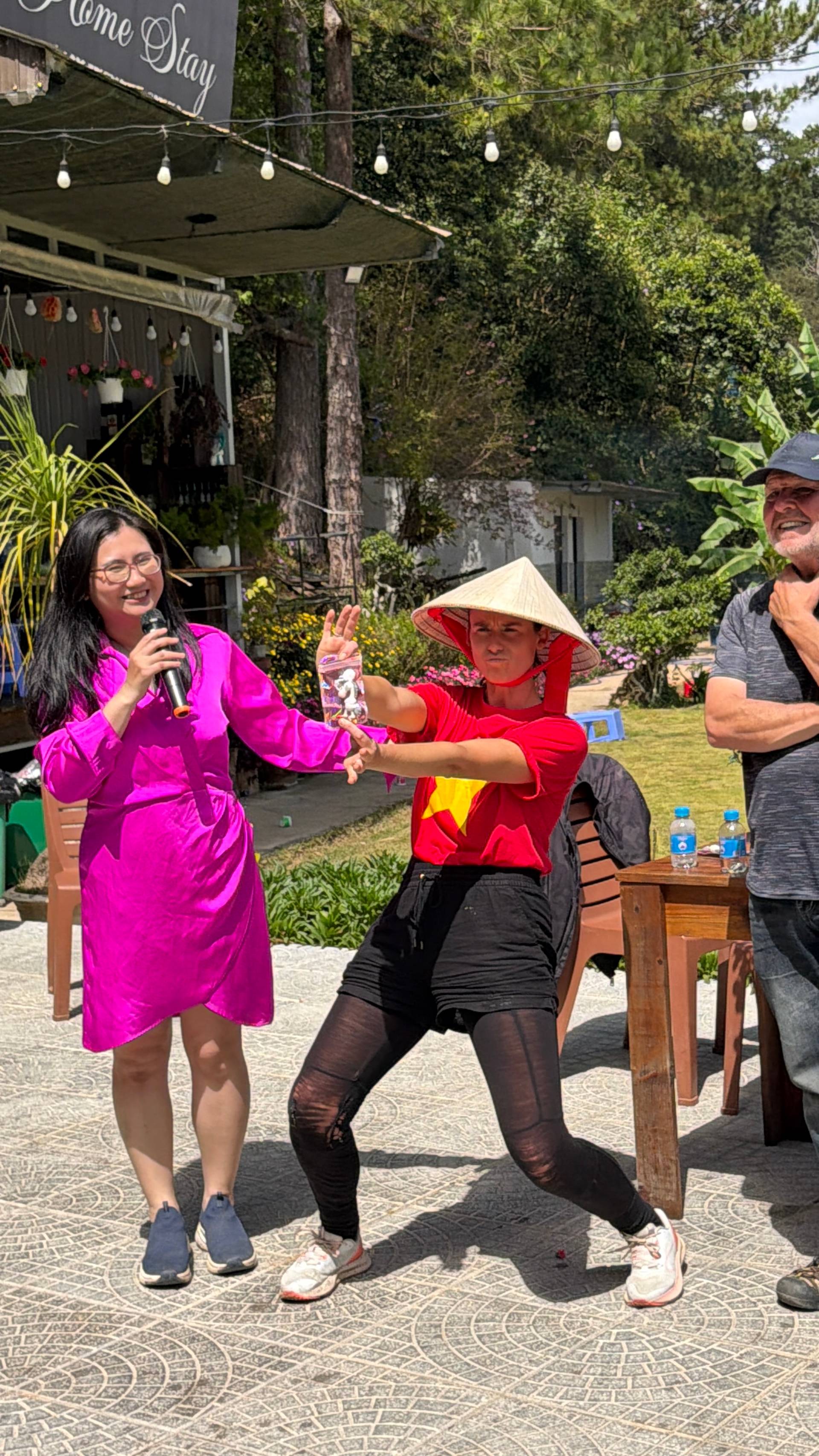 Daytime cultural performance and entertainment event in Đà Lạt featuring colorful traditional costumes, live music with microphone, and interactive audience participation. Set against the backdrop of pine-forested hillsides typical of the region.