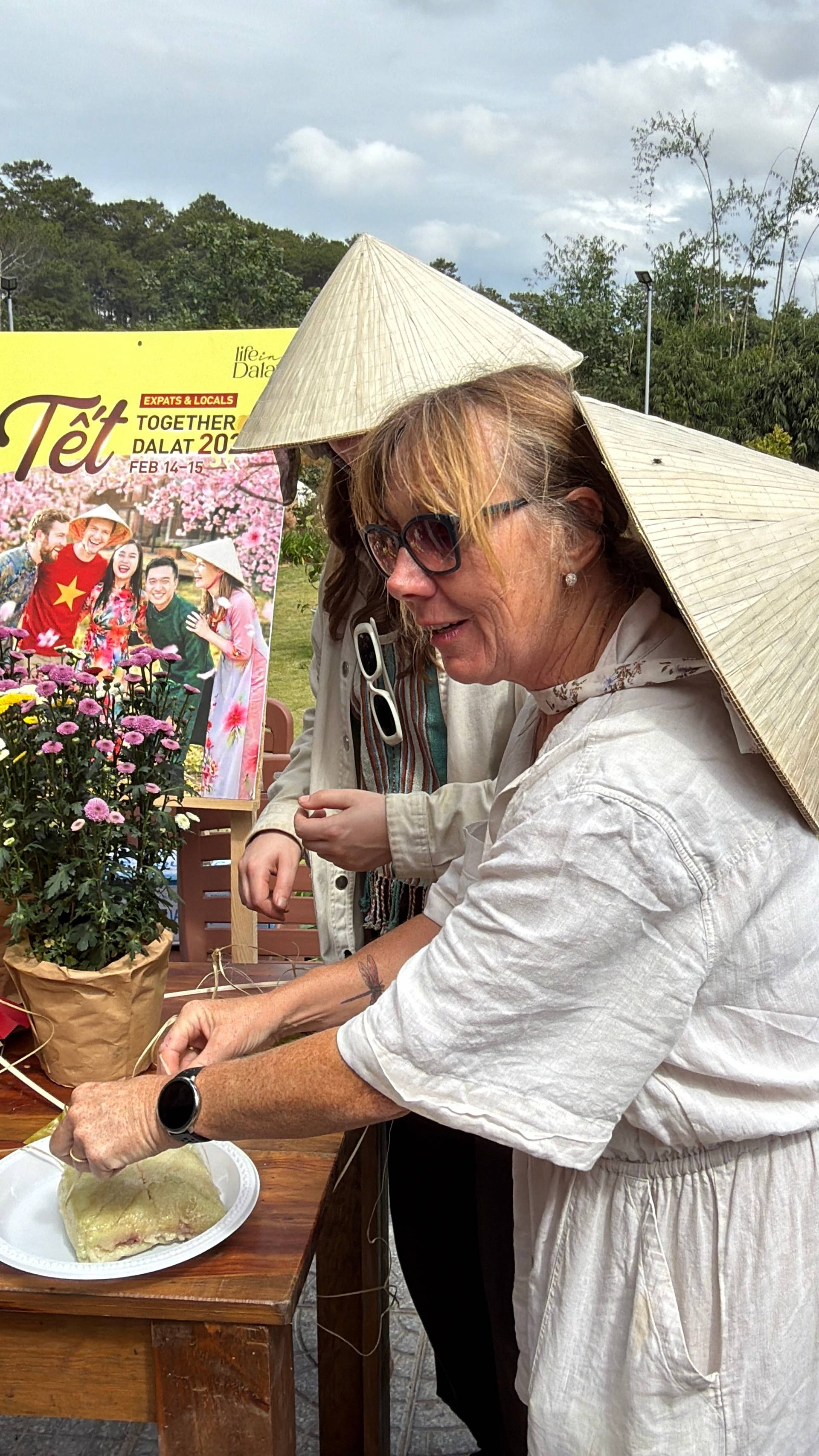Daytime Tết celebration in Đà Lạt featuring traditional Vietnamese cuisine and cultural festivities. Guest enjoying local food under a traditional conical hat with pink flowers and festive banners visible in the background.