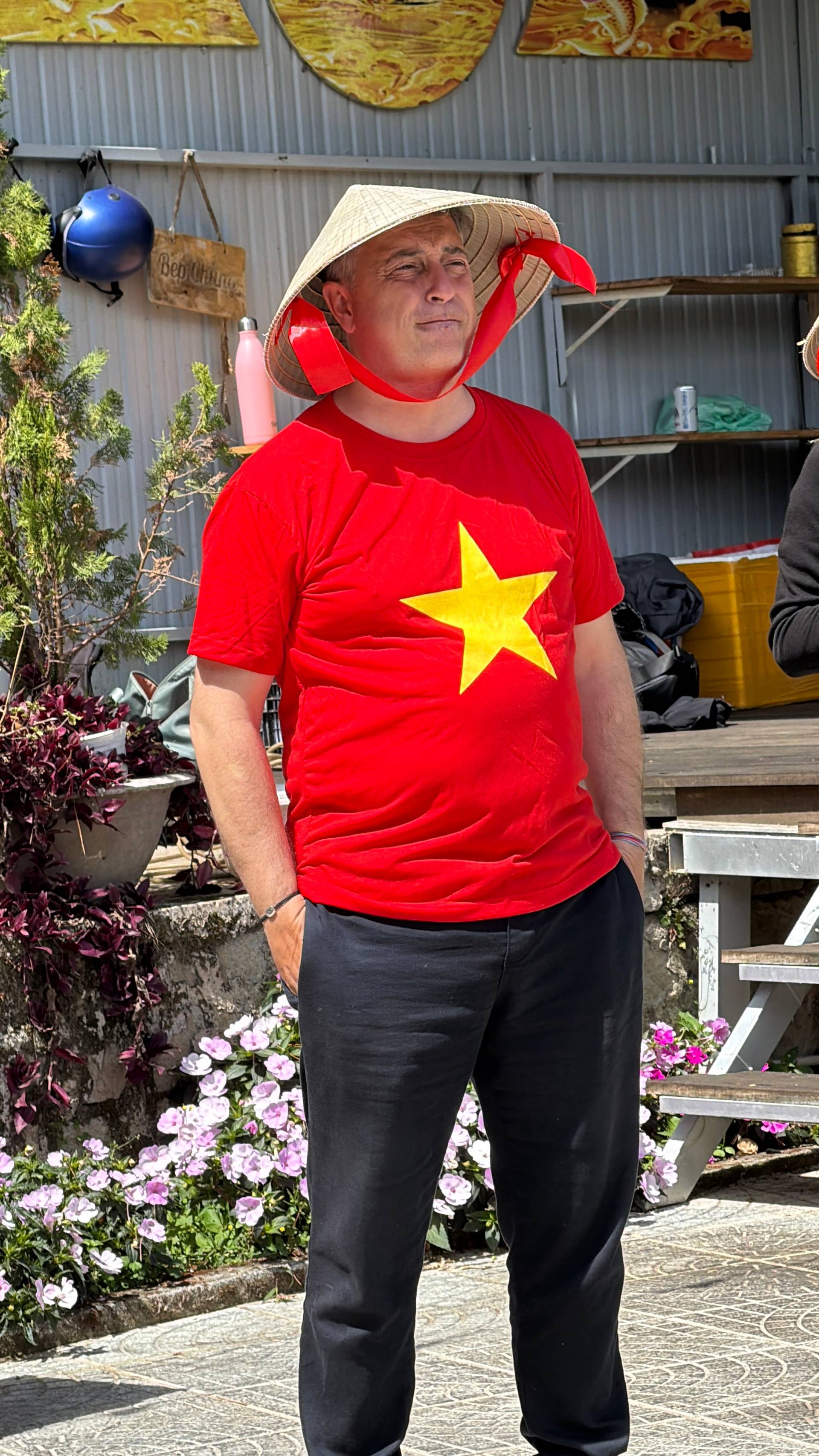 A festival visitor proudly wears the Vietnamese flag on their shirt and a traditional conical hat, standing in a flower-filled garden venue during a Tet celebration in Đà Lạt.