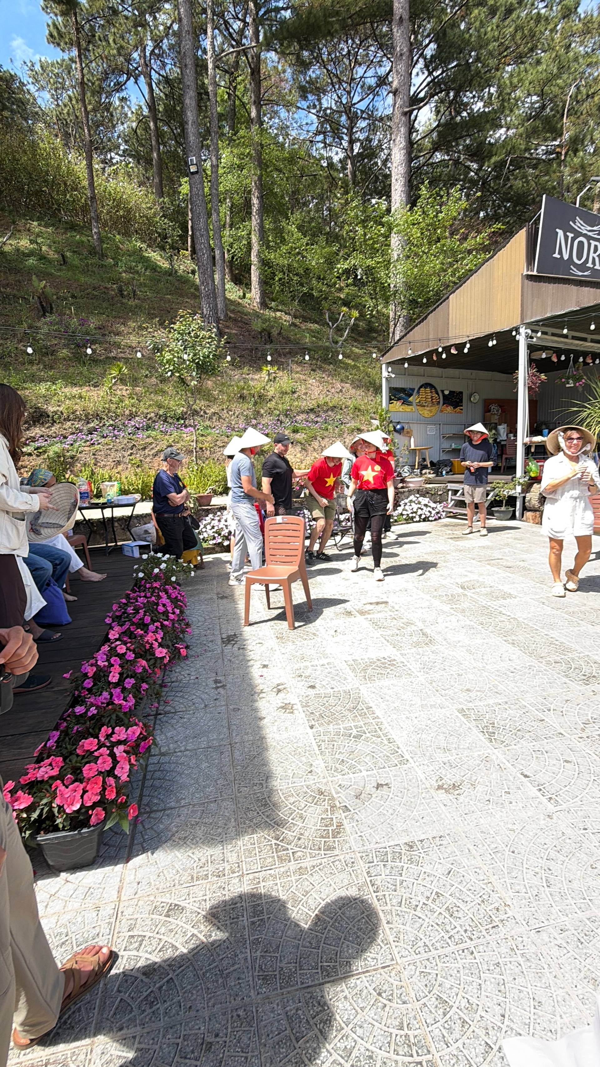 A vibrant outdoor market or festival in a forested hillside setting with visitors wearing traditional Vietnamese conical hats, surrounded by bright pink flowers and a rustic shop venue.