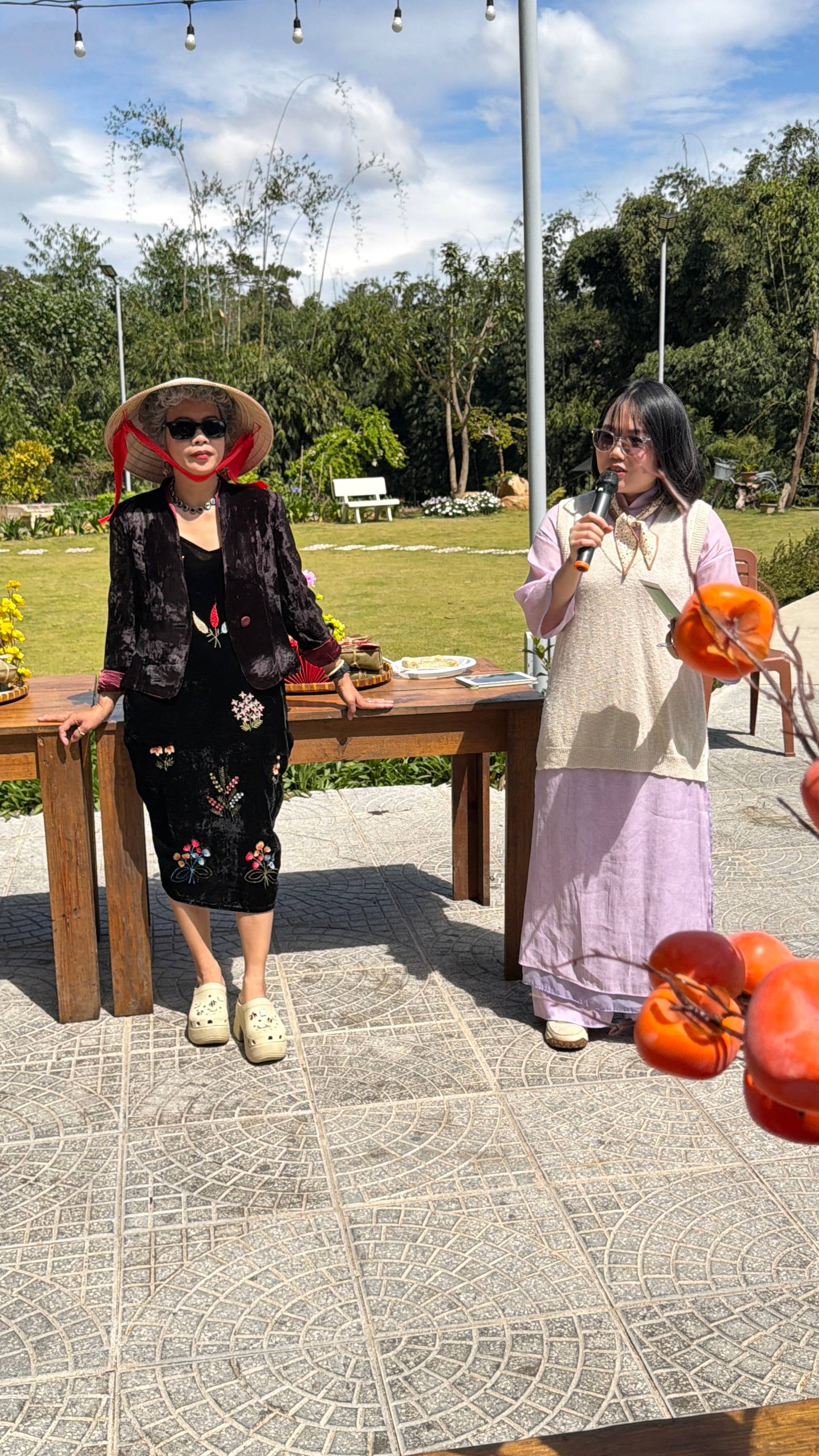 An outdoor event presentation in a lush garden venue with traditional Vietnamese cultural elements, featuring speakers addressing visitors under hanging string lights with pine-forested hills in the background.