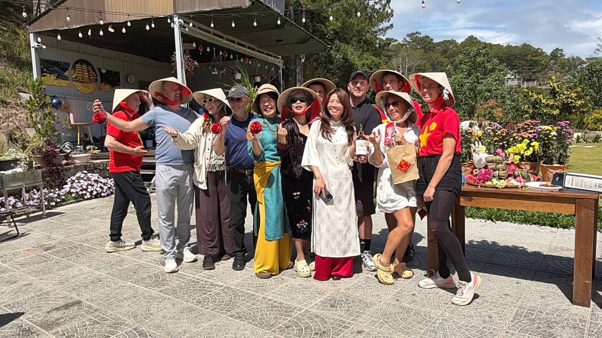 Large group photo of participants at a cultural celebration in Đà Lạt, wearing colorful traditional áo dài and conical hats in a pine-forested outdoor venue.