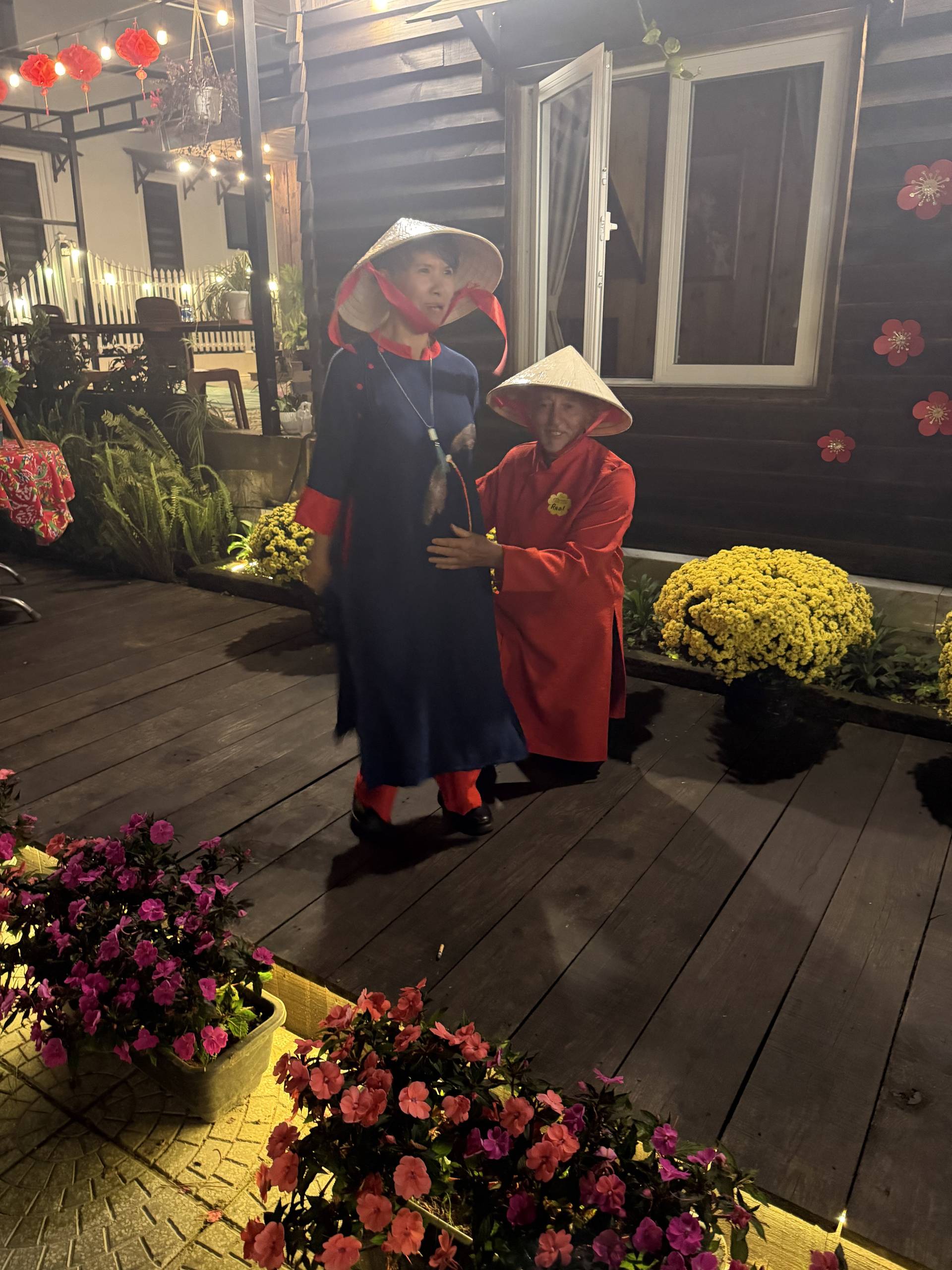 A charming daytime moment in Đà Lạt's highlands featuring visitors in traditional Vietnamese dress posing in front of a decorated wooden residence surrounded by golden chrysanthemums and festival lanterns. The scene captures the warmth of Lunar New Year celebrations in this mountain city's heritage atmosphere.