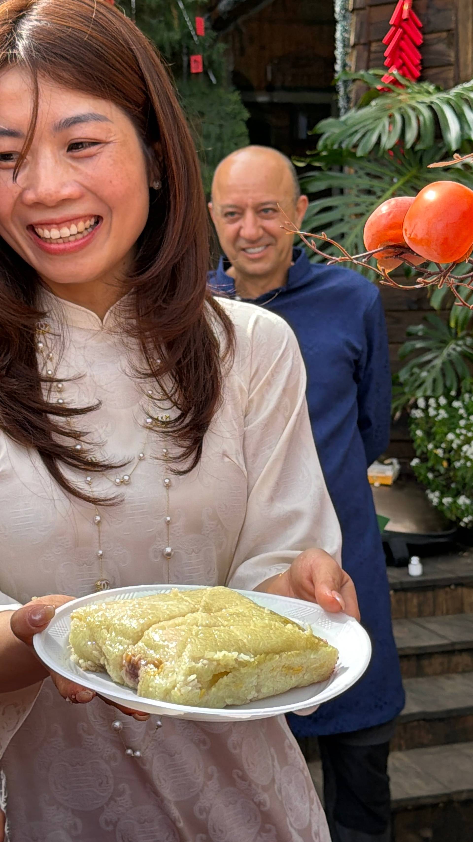 A cheerful Tet celebration moment in Đà Lạt's heritage garden venues captures a visitor in traditional áo dài presenting culinary delights amid vibrant festival decorations and flowering displays. The scene exemplifies the warmth and cultural richness of Lunar New Year celebrations in Vietnam's mountain destination.