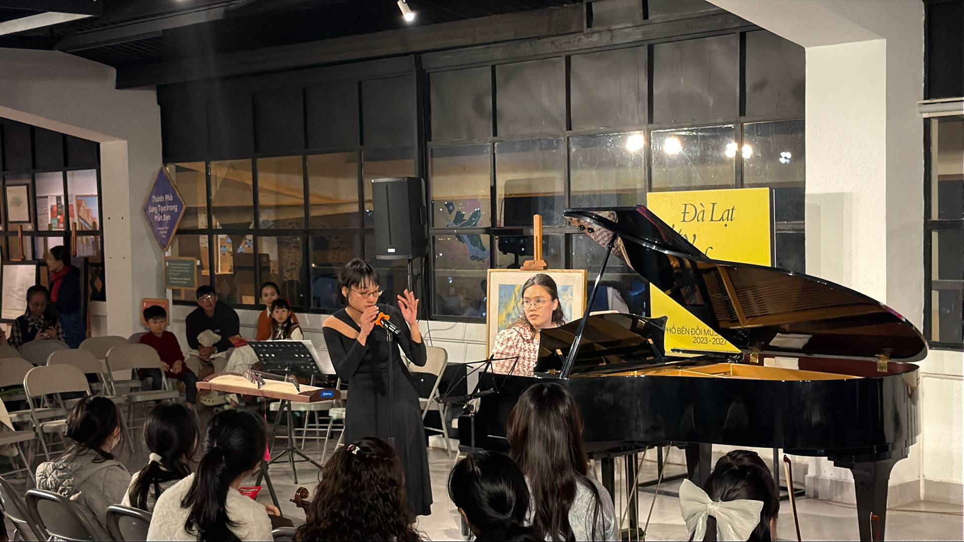 A live classical music event in Đà Lạt's contemporary gallery captures a speaker and pianist performing before an engaged audience. Attendees document the cultural program, reflecting community enthusiasm for classical music and artistic heritage.